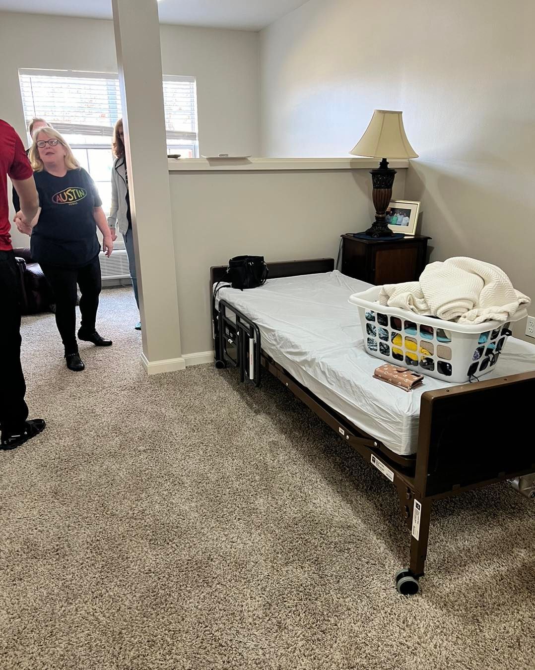Hospital bed in a room, laundry basket on bed, nightstand with lamp. People in background.
