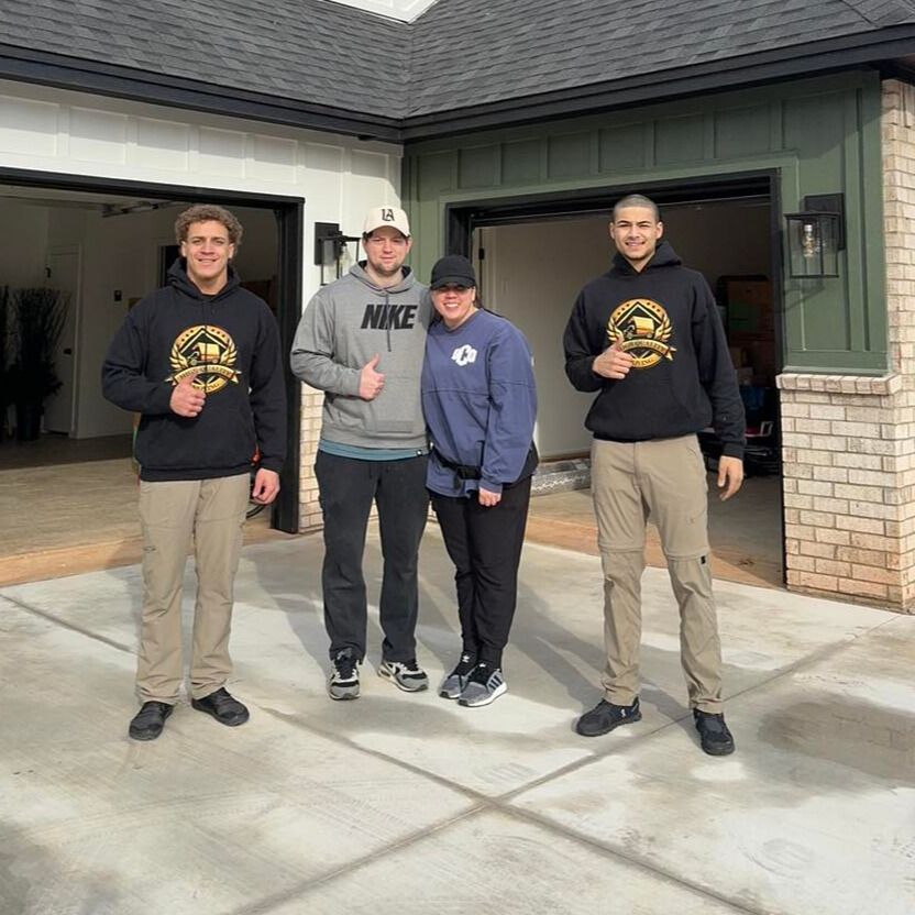 Four people standing in front of a house with a garage; two in black sweatshirts and khaki pants, others in casual wear.