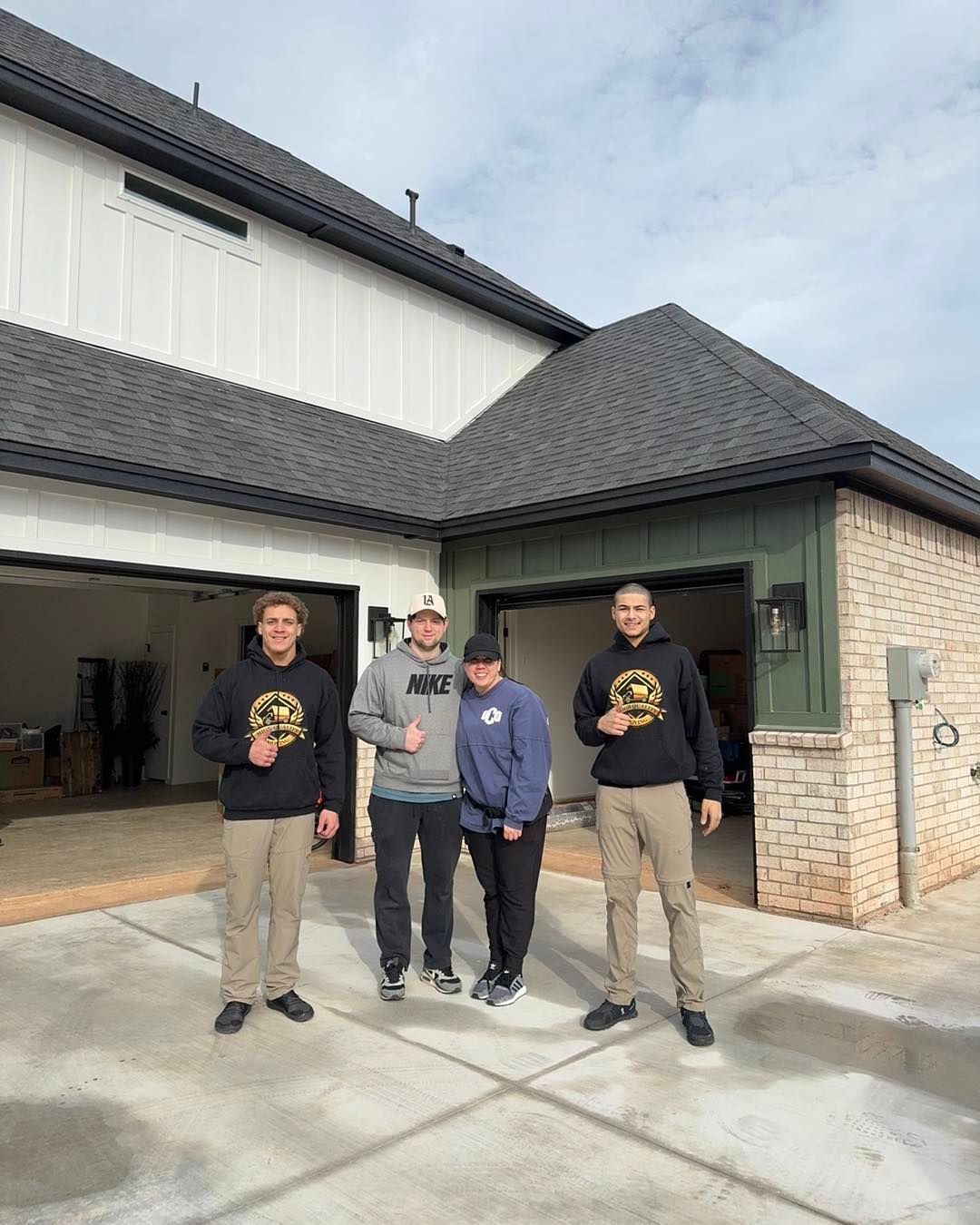Four people stand in front of a house with a garage. They wear casual clothing and hold small objects, possibly keys.