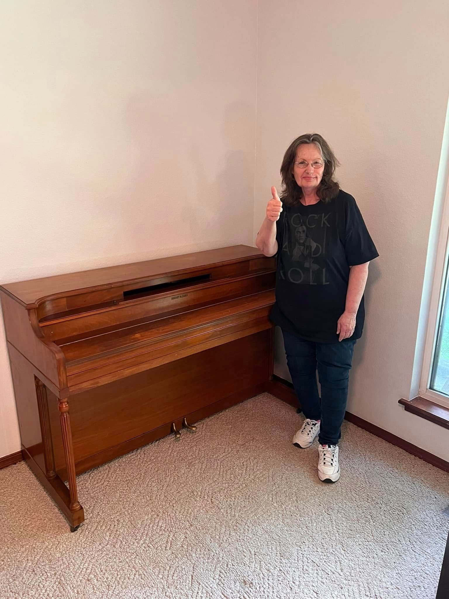 Woman giving thumbs-up next to a wooden upright piano in a room with a window.