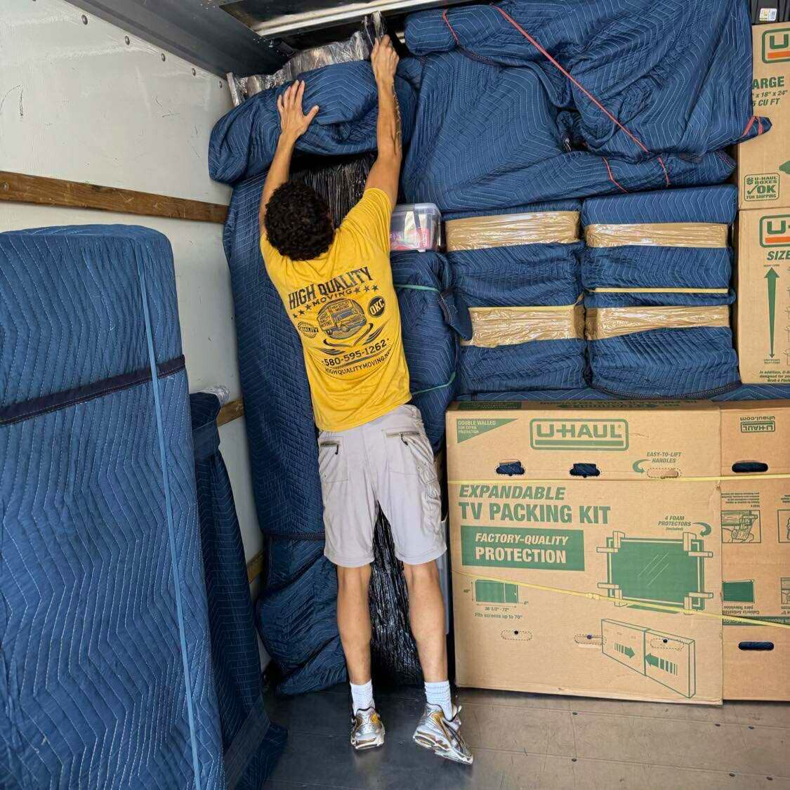 Person in a yellow shirt reaching for something in a packed moving truck. Boxes and furniture pads visible.