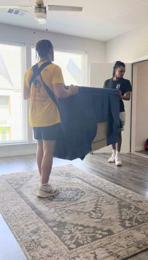 Two people carry a dresser across a room, using straps. They stand on a rug in an apartment.