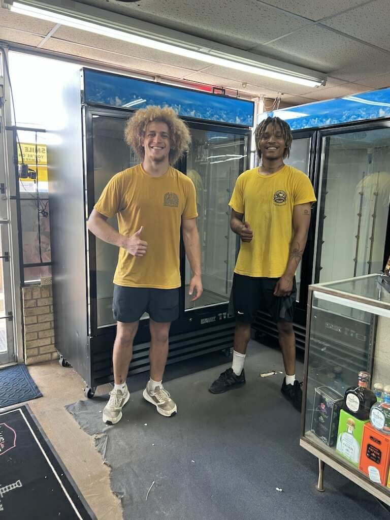 Two people in yellow shirts giving thumbs up in front of a commercial refrigerator.