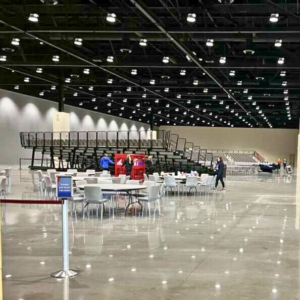 Large event space with tables, chairs, bleachers, and high ceiling with many lights. People are present.