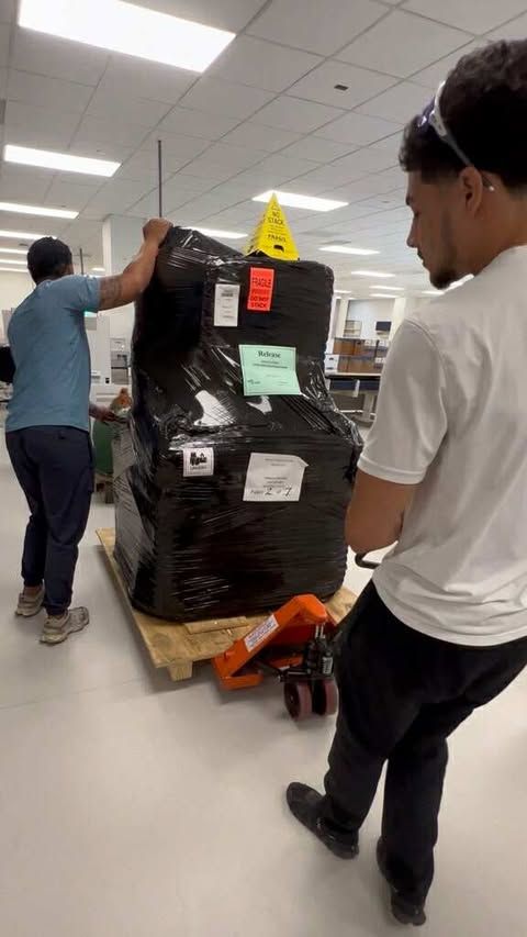 Two movers moving a large, wrapped pallet with a hand truck in a hospital