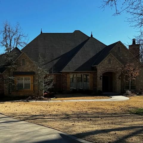 A large, brick home with dark roof on a sunny day. Bare trees and dry grass in the yard.