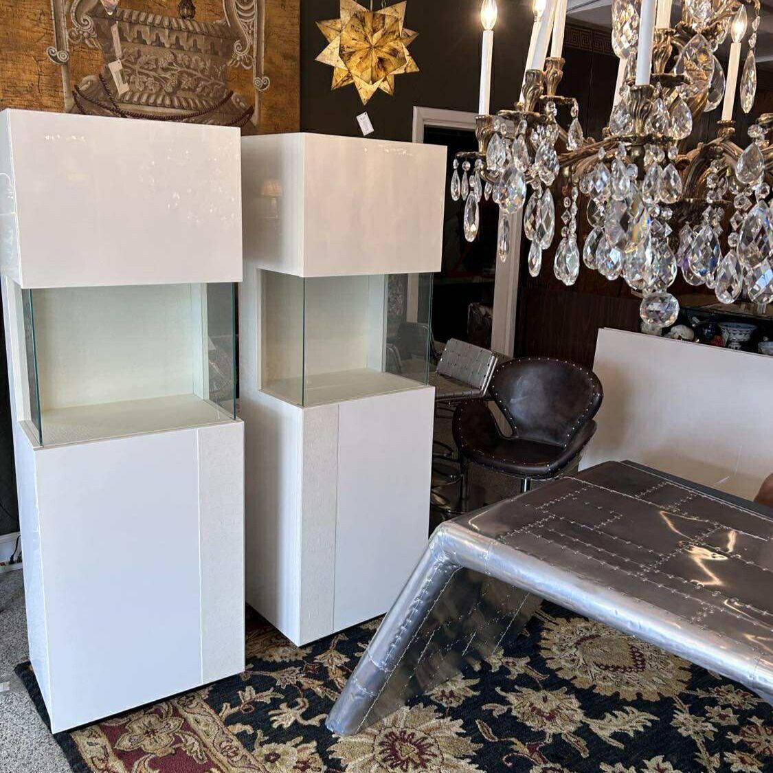 Two white display cases with glass shelves, near a table and chandelier.