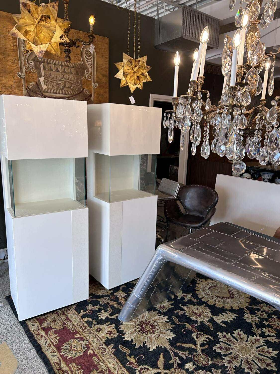 Two white display cabinets with glass shelves, near a chandelier and table.