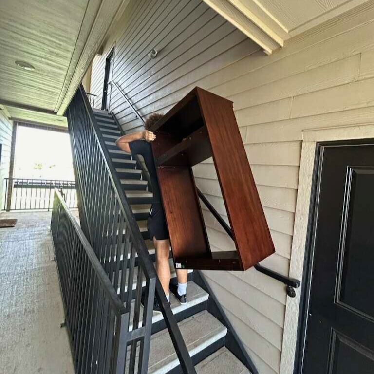 Person carrying a large wooden cabinet up outdoor stairs. Black railings, beige siding, black door.