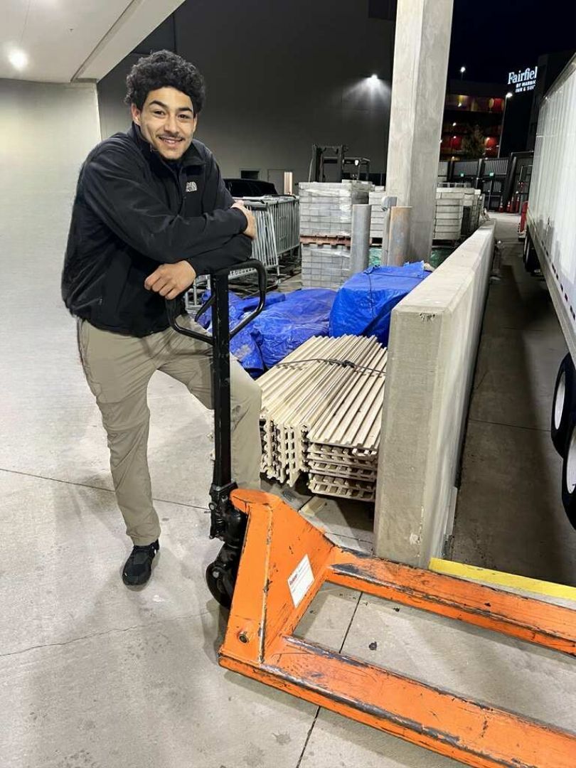 Man leans on a pallet jack at a loading dock. He is wearing a black jacket and tan pants.