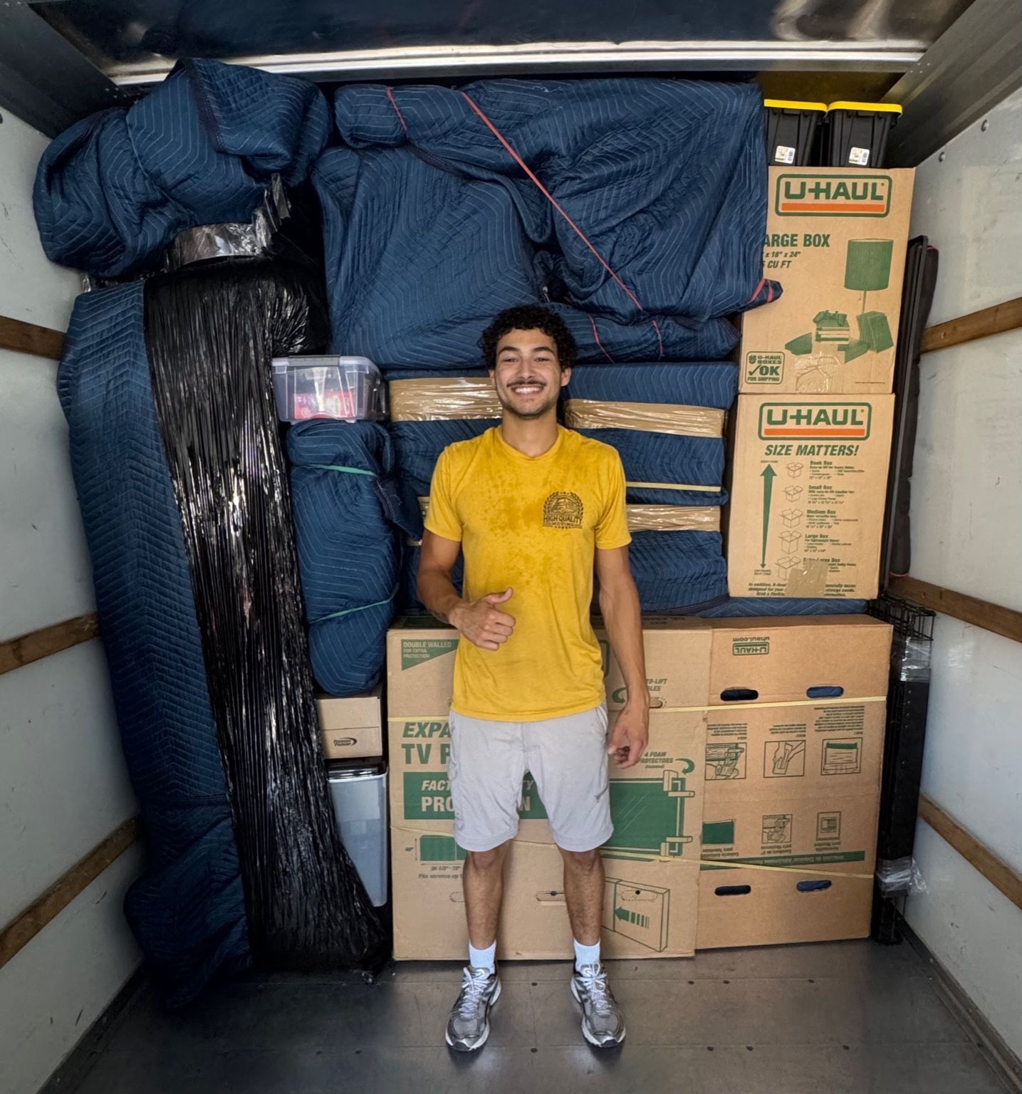 Two men stand in a moving truck filled with packed boxes and furniture. They smile and give thumbs up.