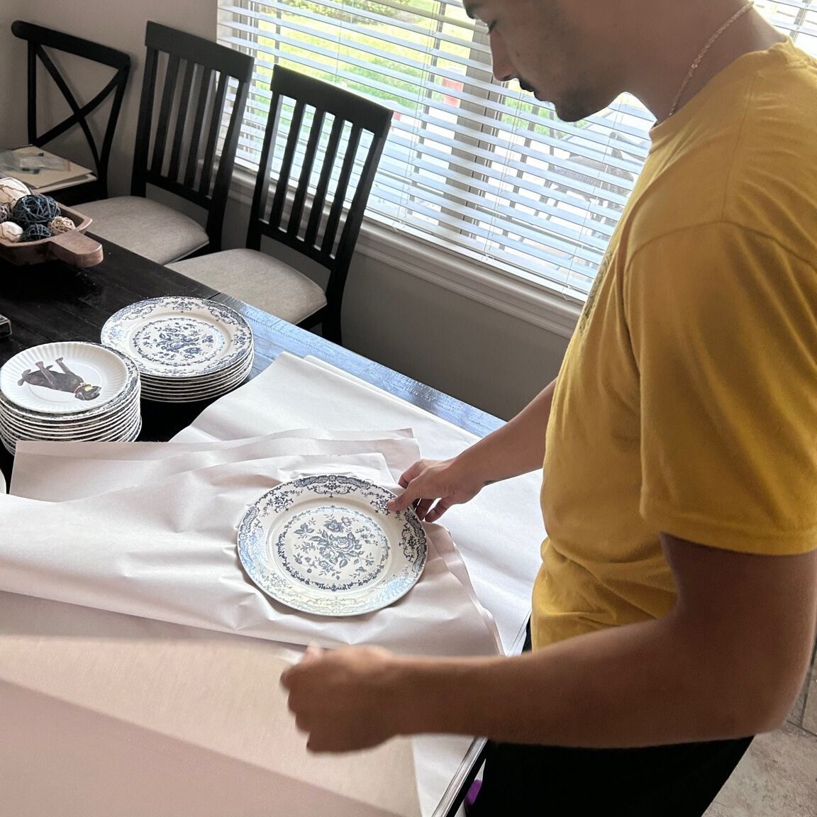 Person wrapping a blue and white patterned plate with white paper on a table, preparing to pack dishes.
