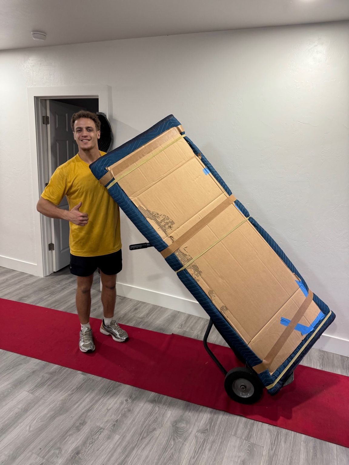 Man moving furniture, wrapped in blue pad, on a hand truck, exiting a garage onto a ramp.