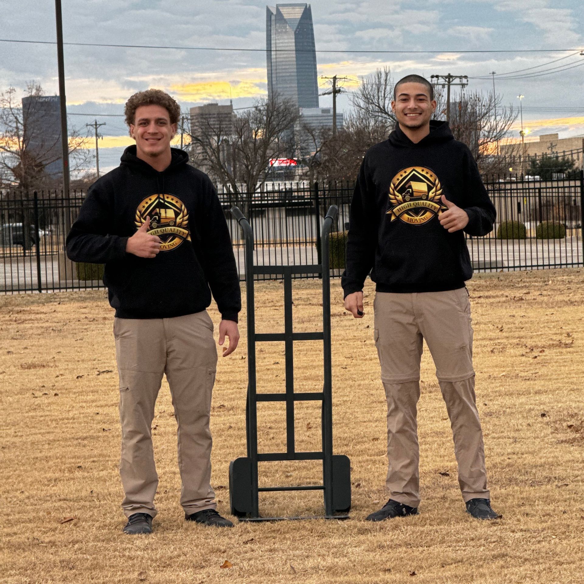 Two men in black hoodies and khaki pants pose with a hand truck outside, with a city skyline in the background.