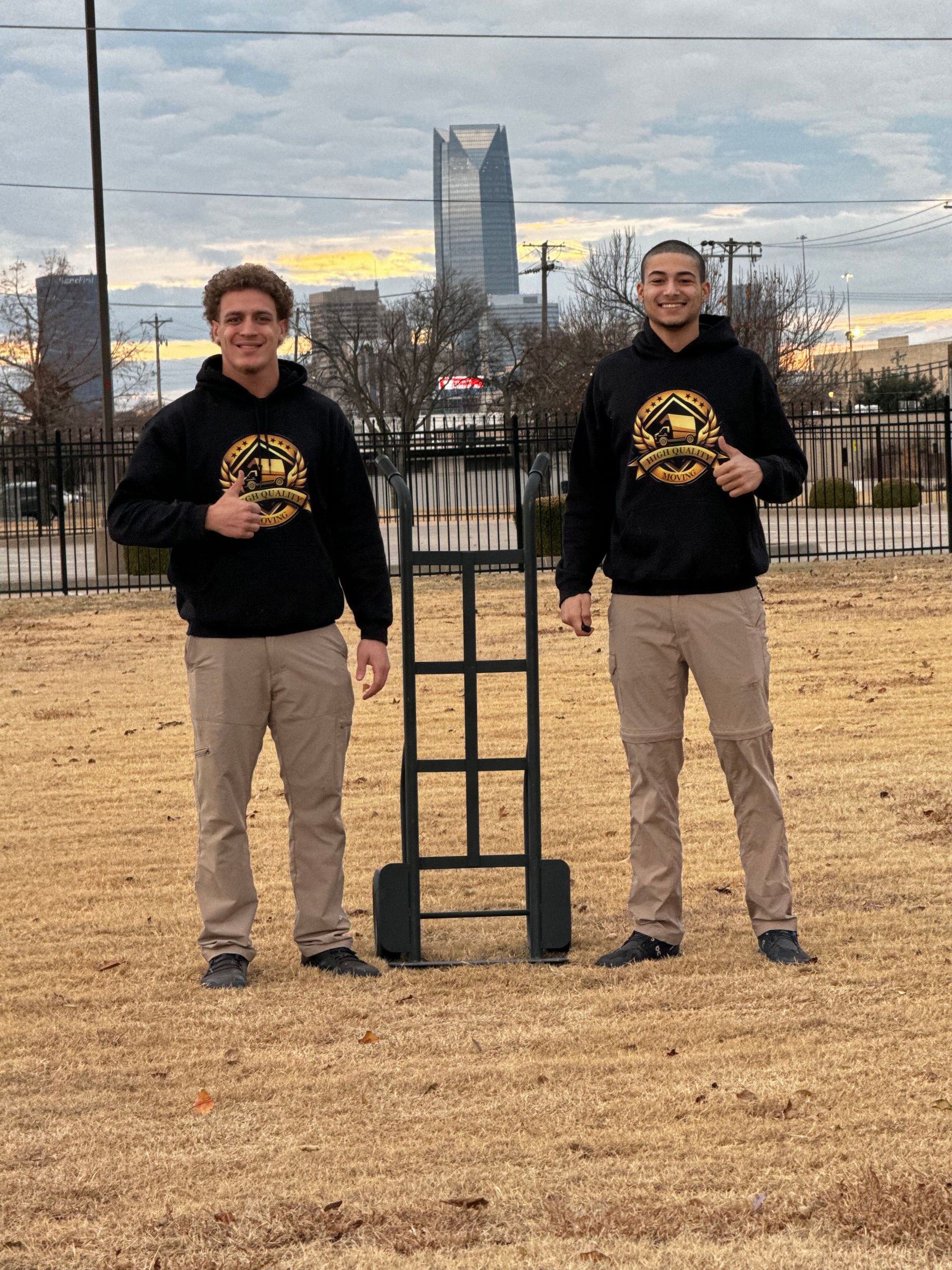 Two men in black hoodies and khaki pants stand with a hand truck outside, OKC skyline in the background giving thumbs up.