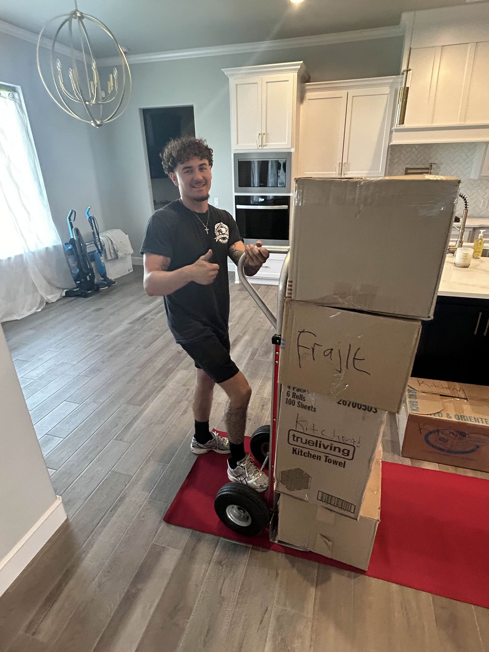 Man with thumbs-up pushing boxes on a dolly inside a home with hardwood floors and kitchen.
