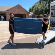 Two men carrying a large blue padded item from a moving truck in front of a house on a sunny day.