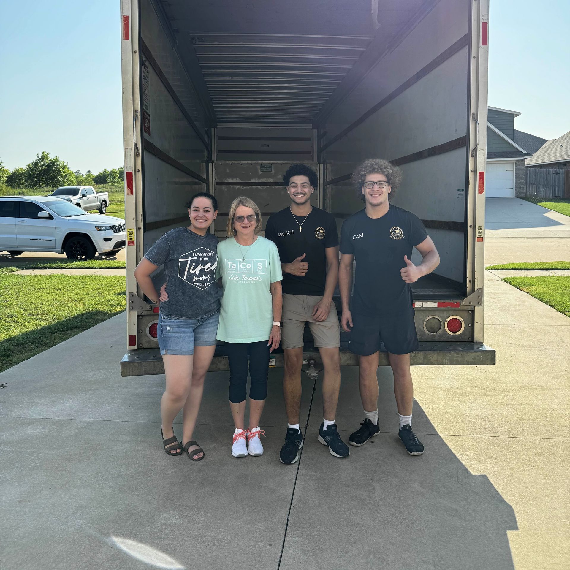Four people stand in front of an open moving truck. Two men give thumbs up. Sunny outdoors.