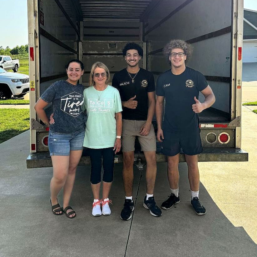 Four people smiling, posing in front of an open moving truck. People in casual clothes on a sunny day.