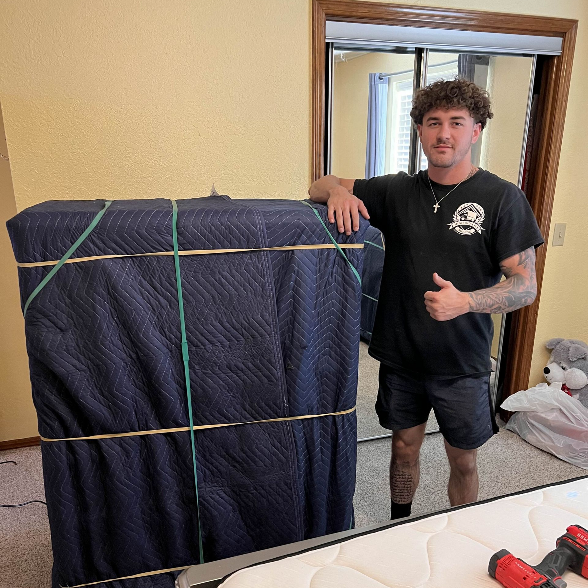 Man with thumbs-up next to a large, wrapped, secured object in a room.