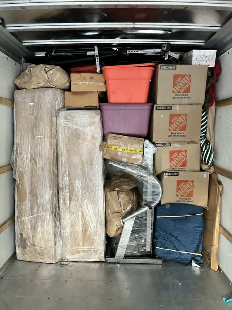 Packed moving truck interior with furniture wrapped in plastic, boxes, and bins.