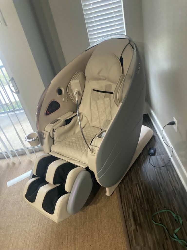 White and gray massage chair indoors, reclined, with cup holder and foot massager, next to a wall and window.