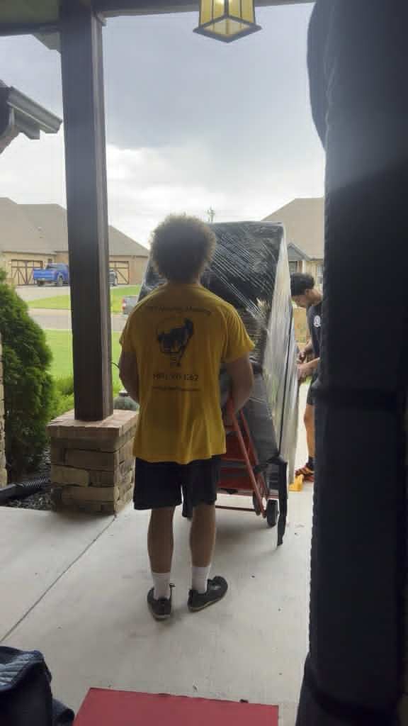 Movers carrying a large, wrapped gun safe on a hand truck out of a house under a porch, on a cloudy day.