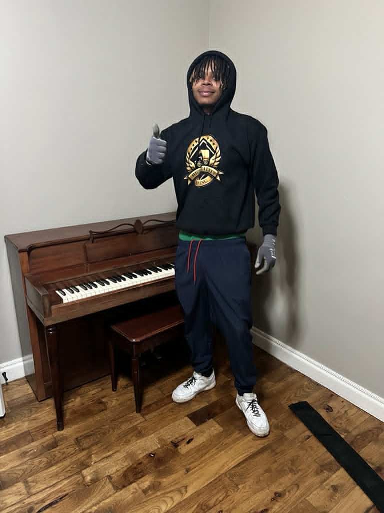 Man in black hoodie gives thumbs up near a piano in a room with light-colored walls. He wears gloves.