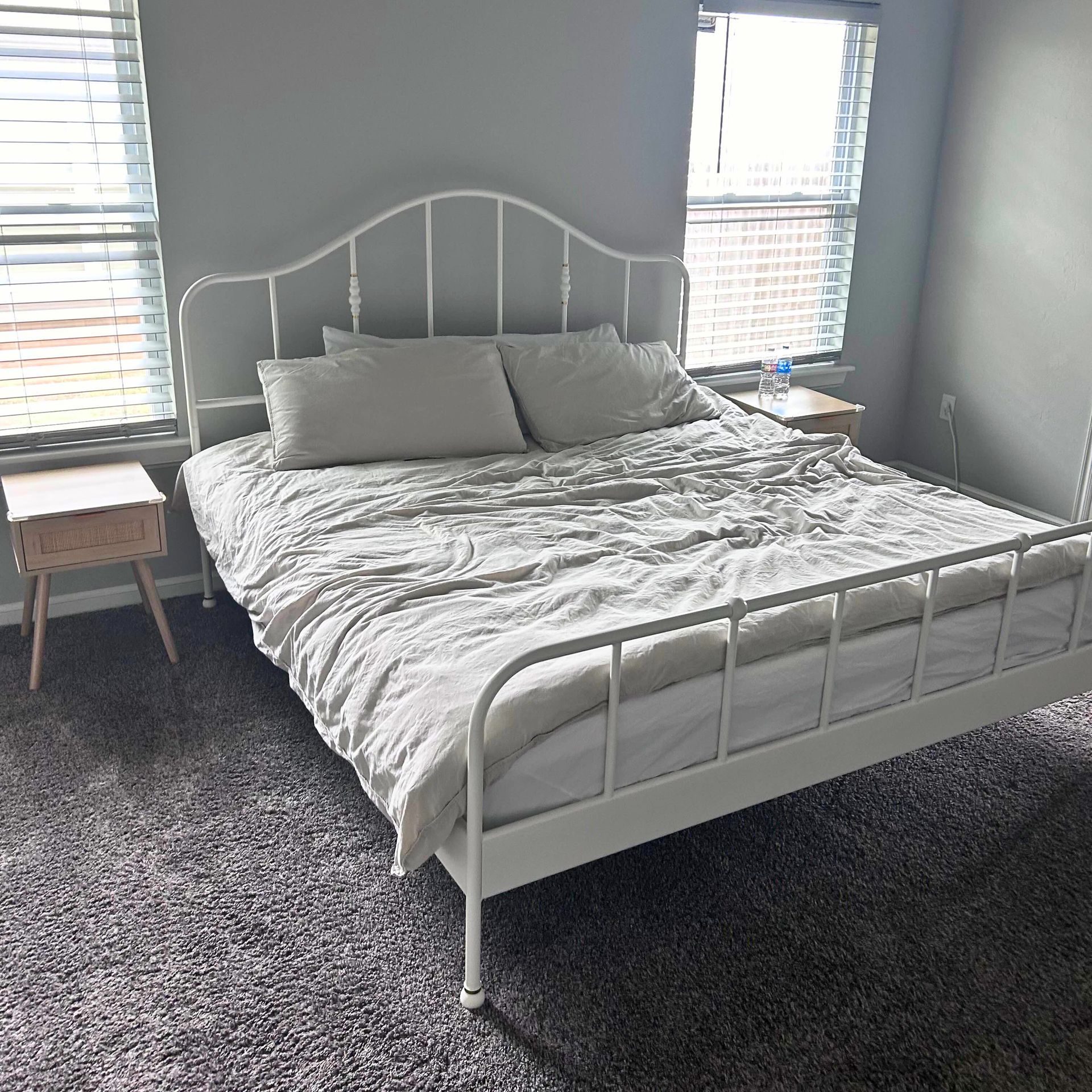 White metal bed with gray bedding, flanked by two wooden nightstands, between two windows.