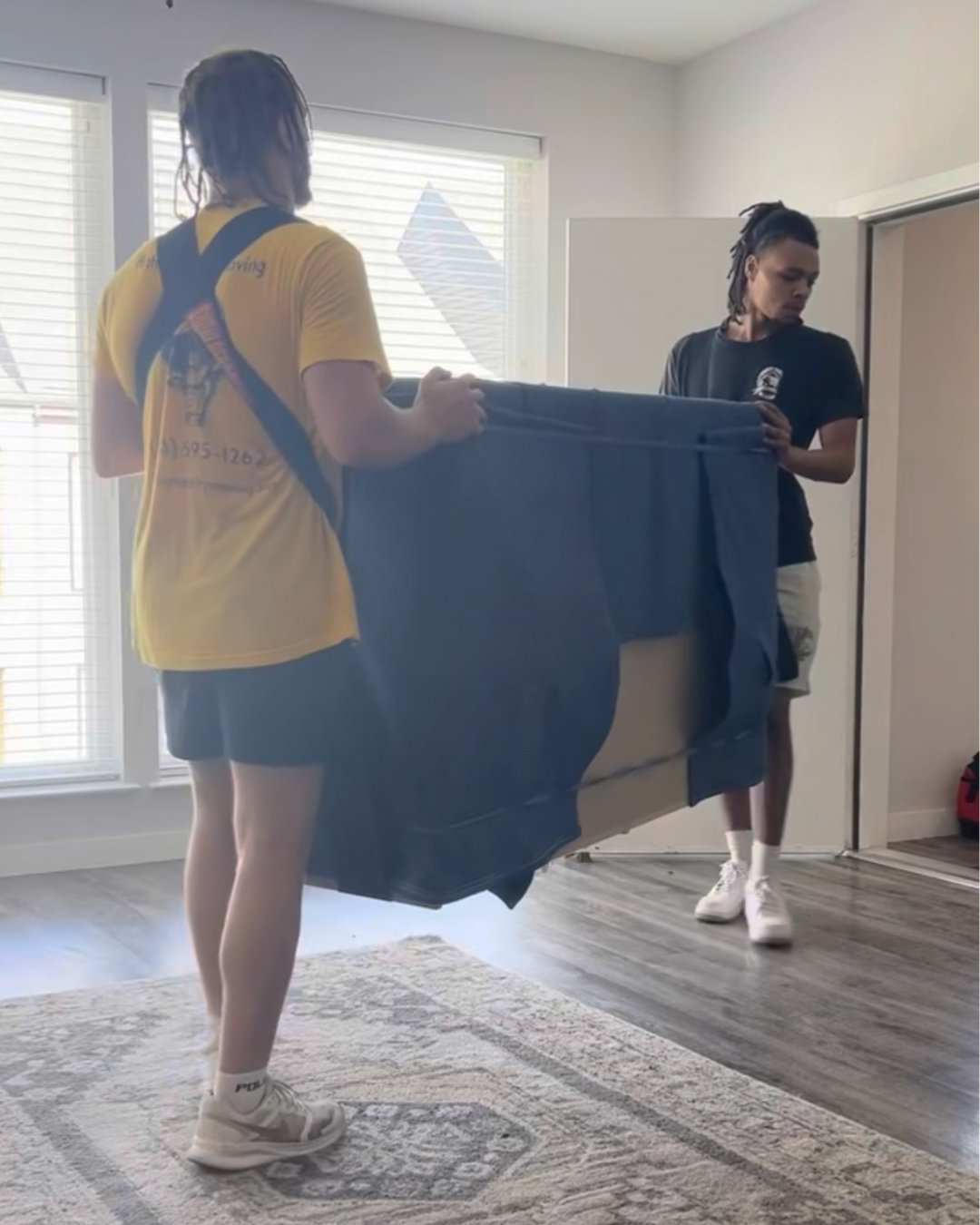 Two people carrying a large, blue couch through a room with a rug and window.