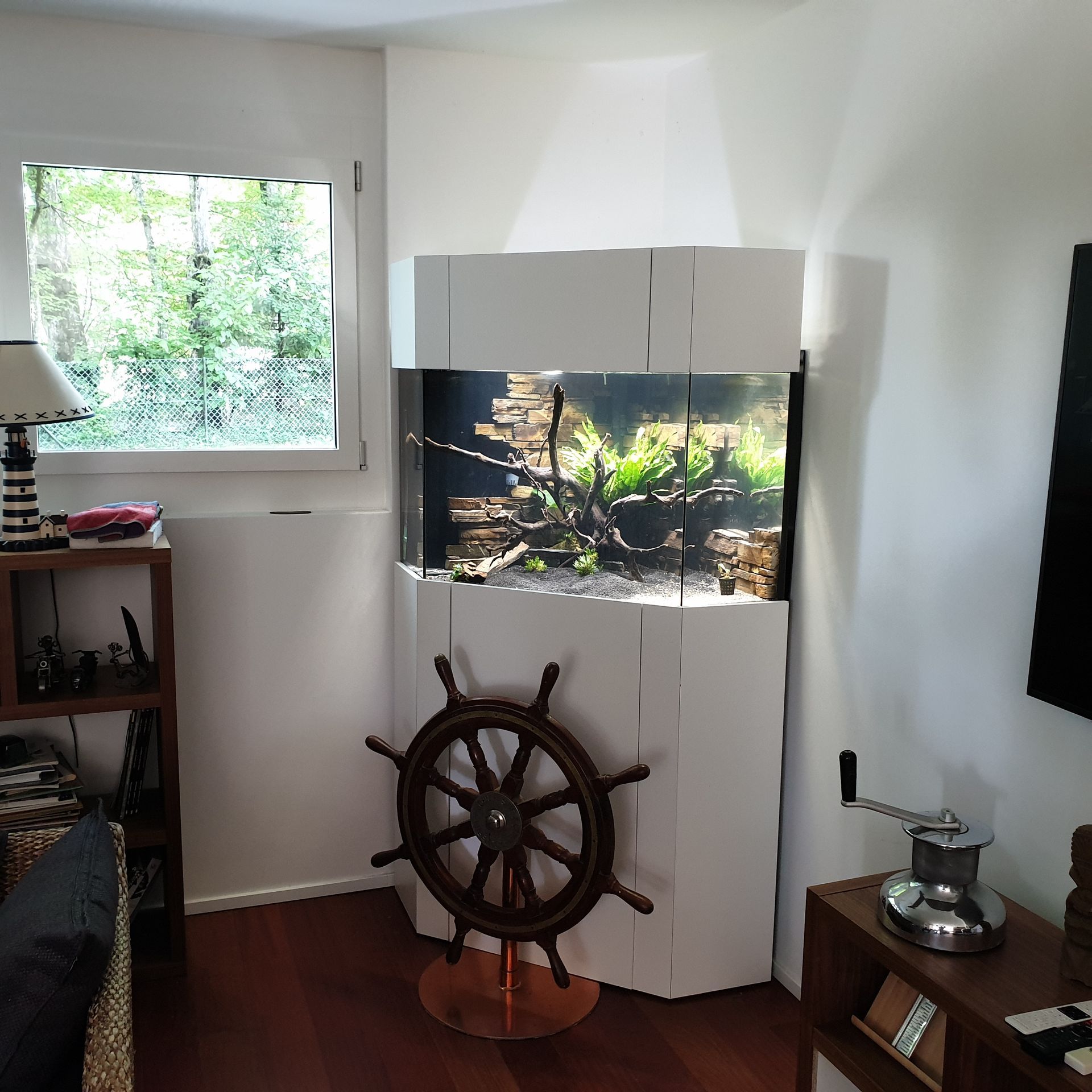 Aquarium d'angle avec meuble blanc, à côté d'une fenêtre et d'une barre de bateau.