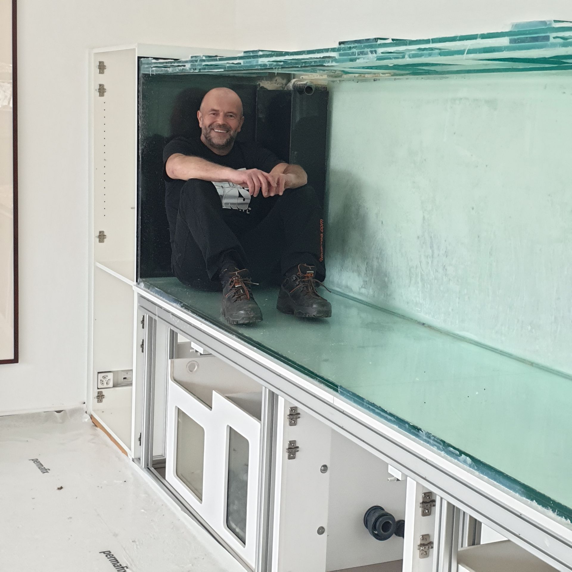 Un homme, souriant, est assis à l'intérieur d'un aquarium. Vêtu de noir, il est assis au-dessus d'un bac de décantation dans une pièce blanche.