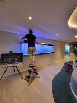 Une personne sur un escabeau ajuste un aquarium fixé au mur dans une pièce au parquet clair et à l'éclairage bleu et blanc.