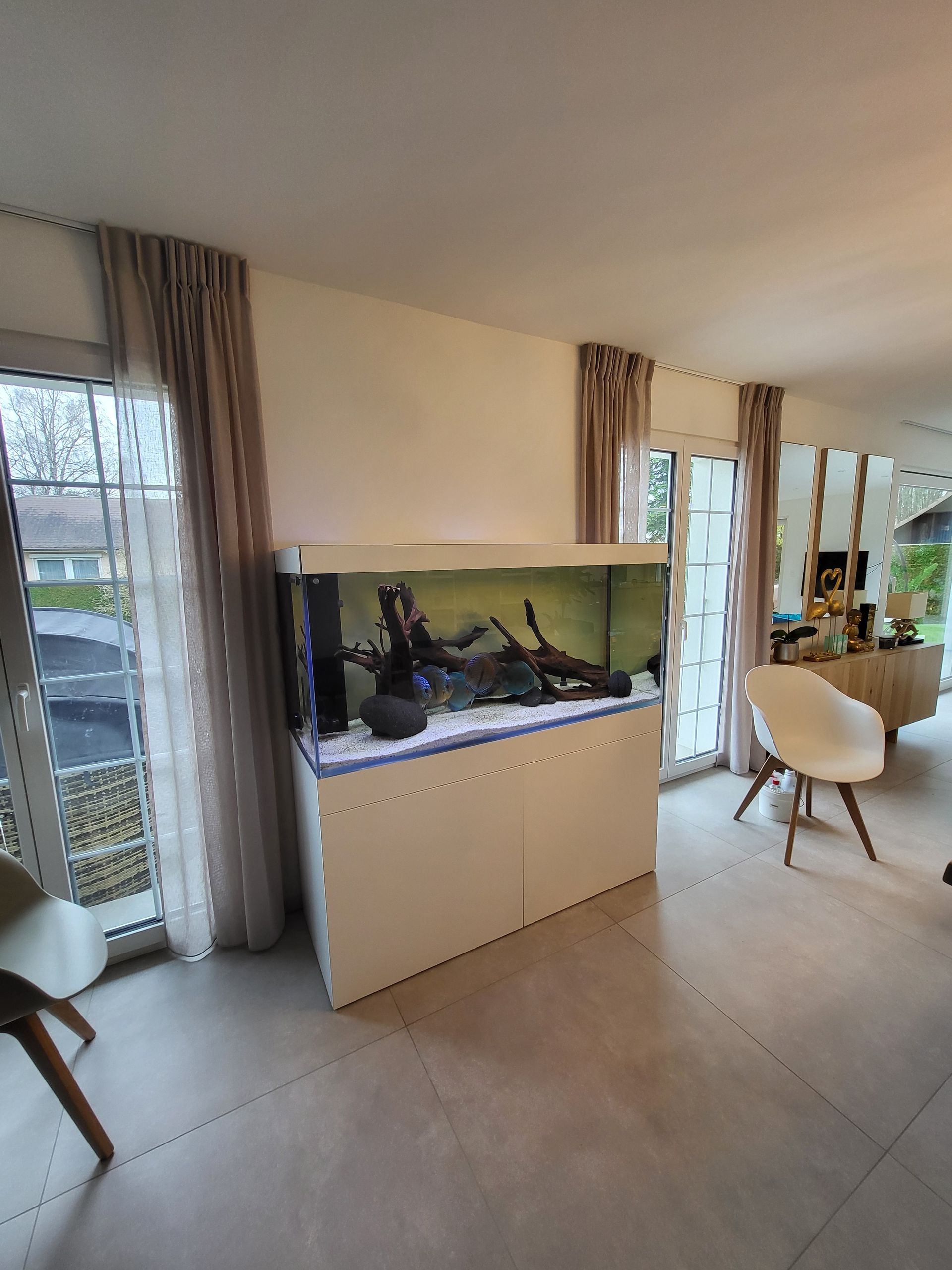 Un grand aquarium rectangulaire avec du bois flotté et des pierres dans un meuble blanc contre un mur blanc dans un salon.