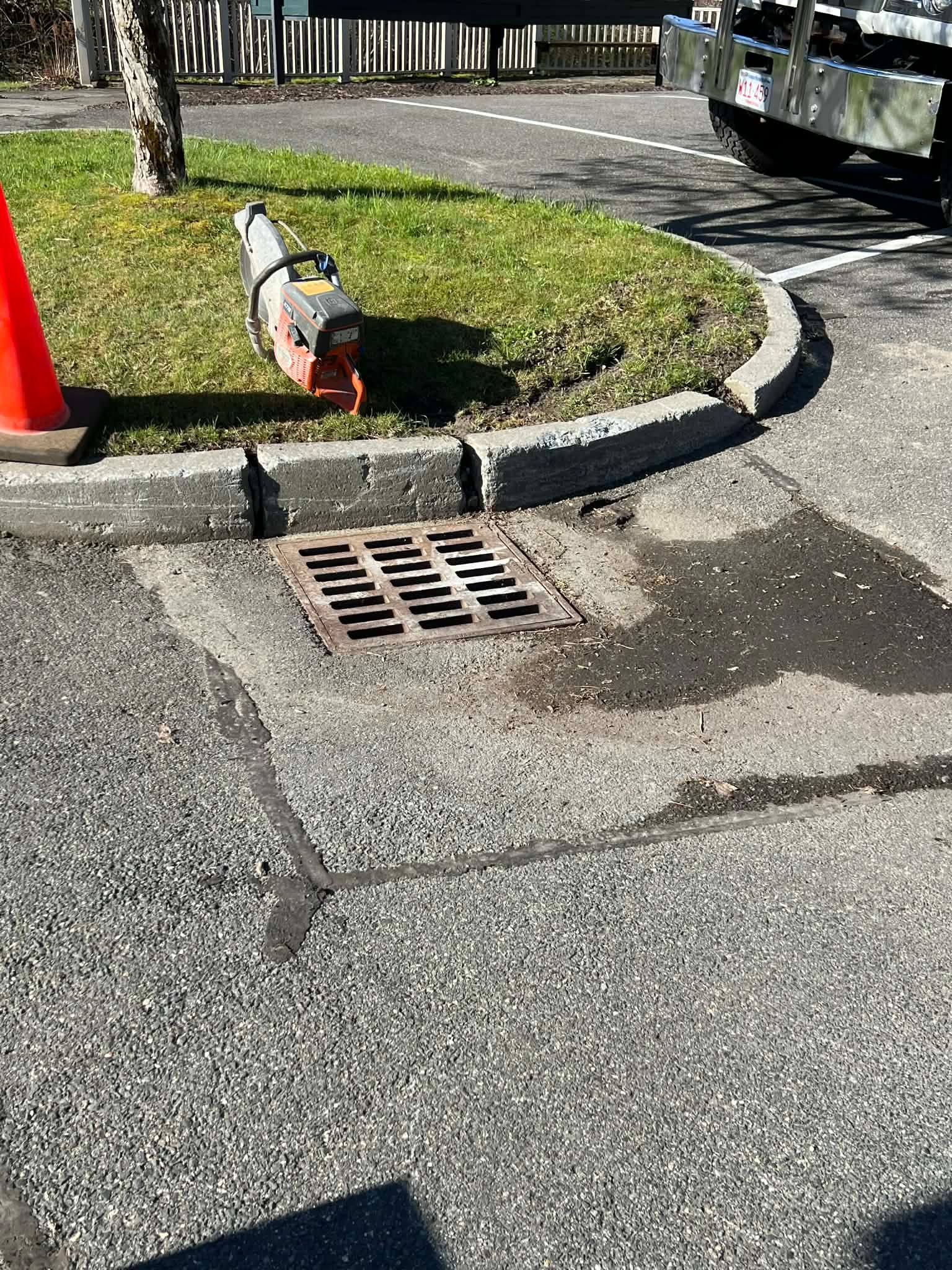 Drainage grate on asphalt curb; saw on grass; orange traffic cone.