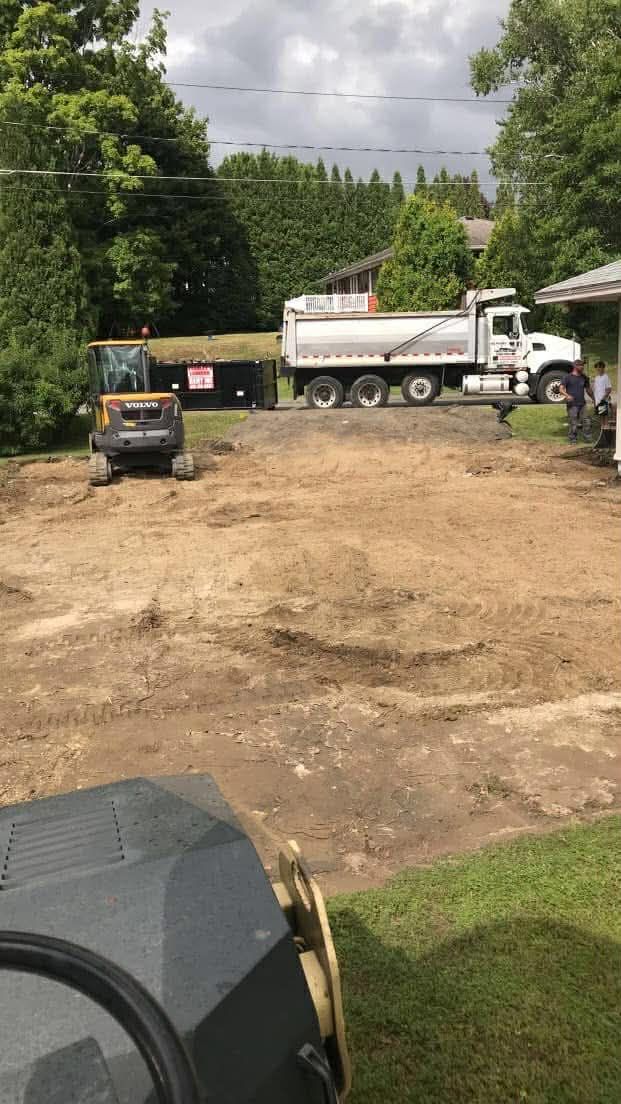 A construction site with a truck, excavator, and leveled dirt. Green trees and a house are in the background.