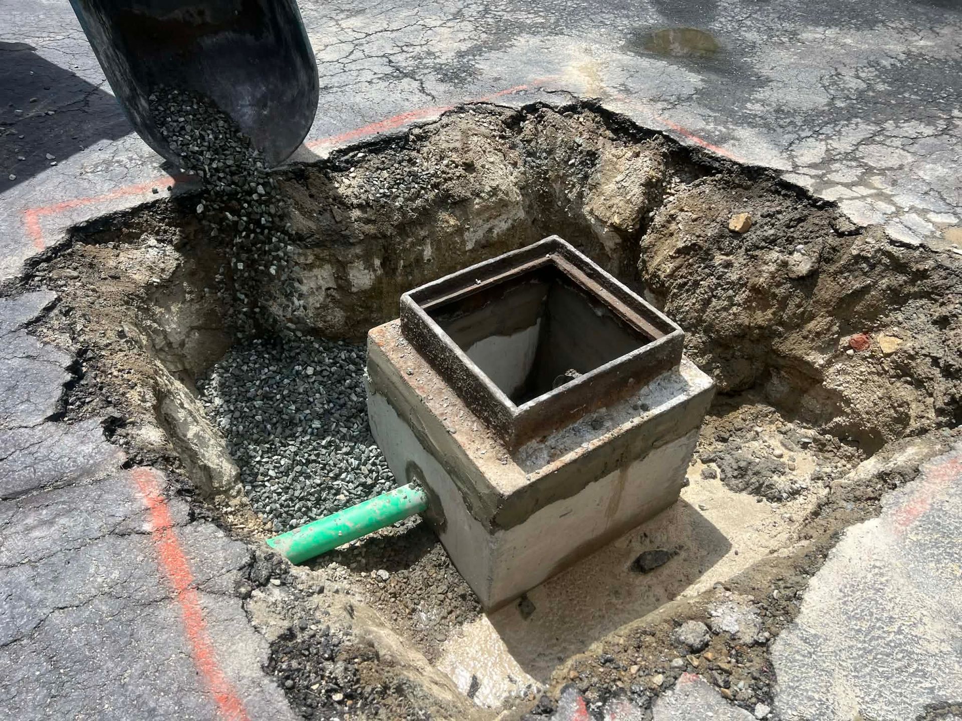 A utility box surrounded by asphalt in a road, being filled with gravel from a black container.