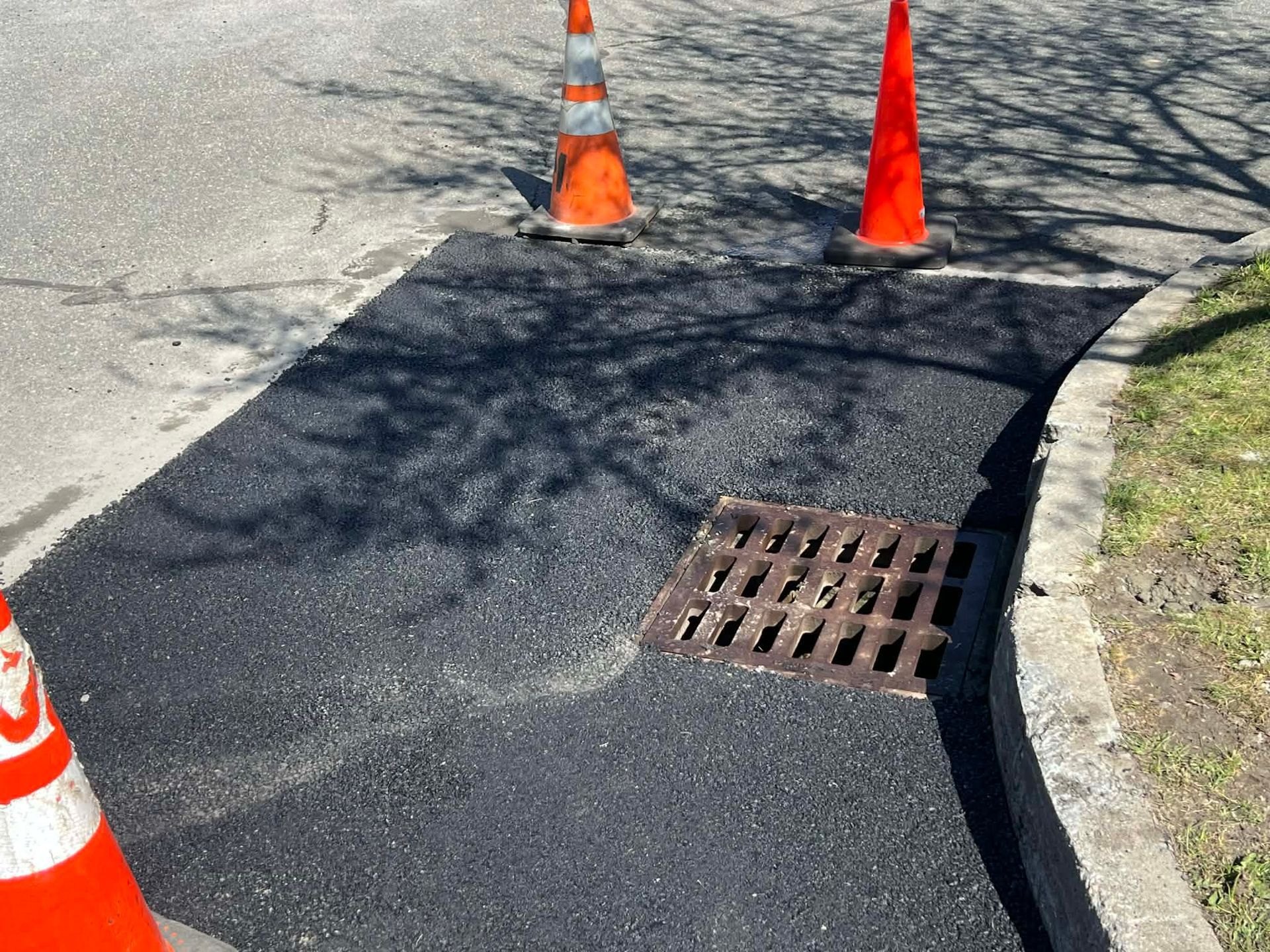 Newly paved asphalt patch with drainage grate, marked by orange traffic cones.