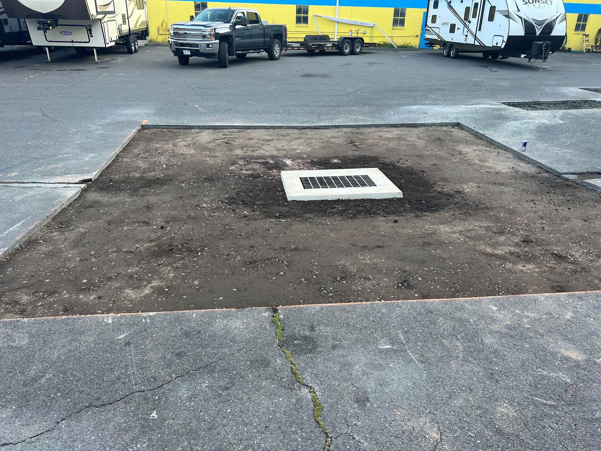 Asphalt square with inset drain, surrounded by dark soil, in a parking lot.