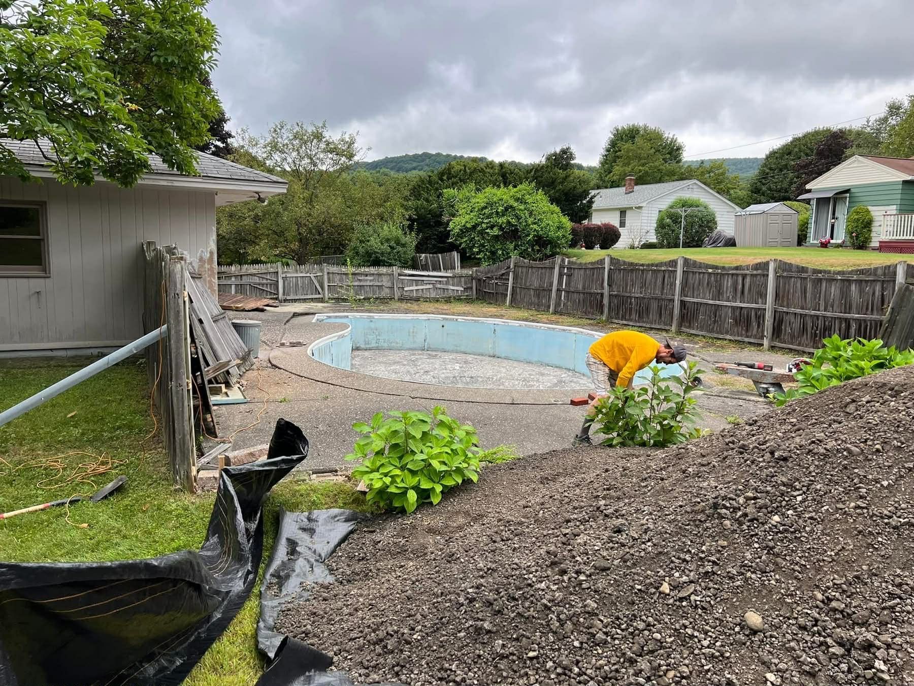 A person works near an empty, in-ground pool surrounded by a fence and houses.