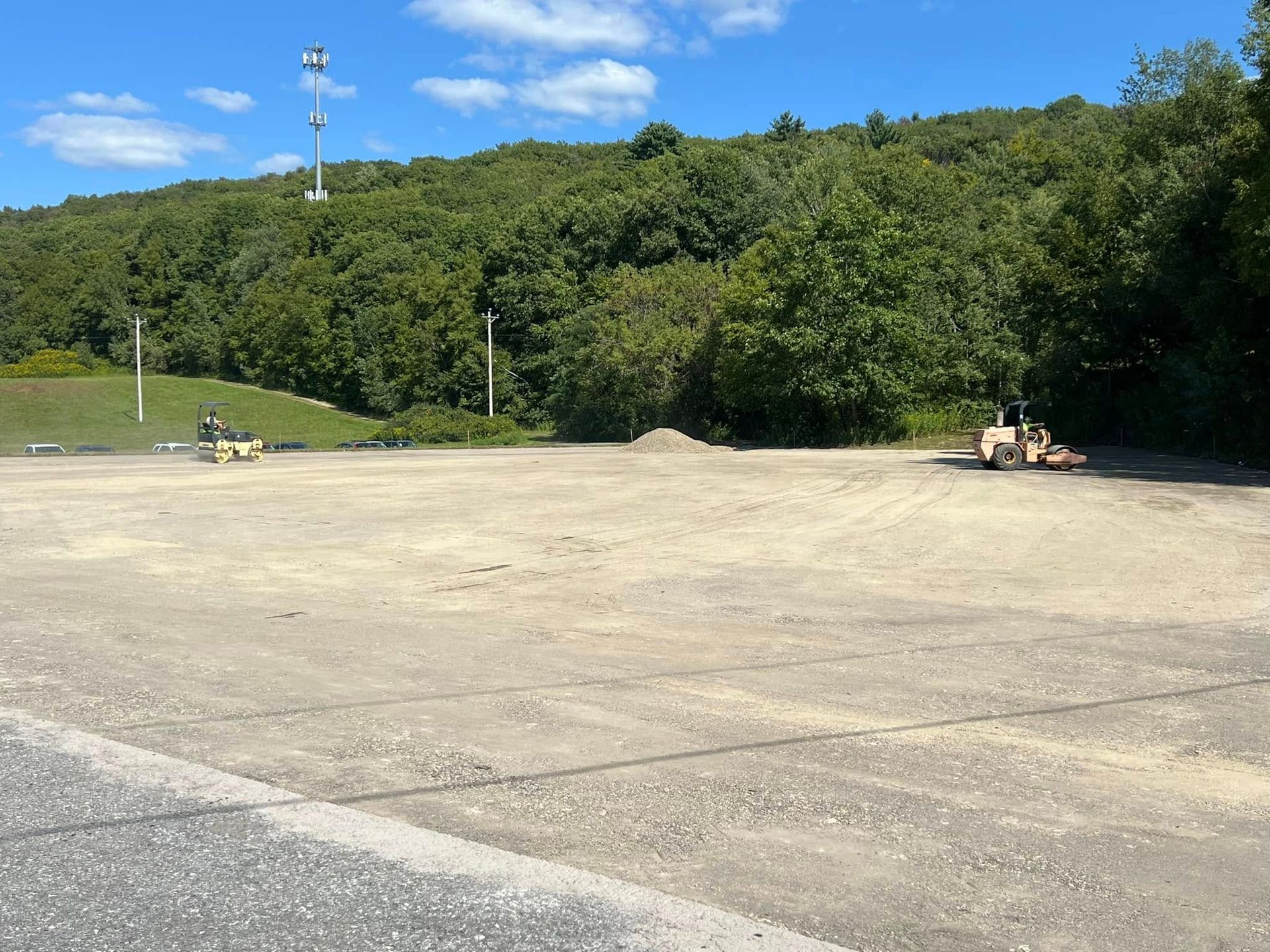 Two small yellow construction vehicles compacting dirt in a clearing with a treed hill in the background.