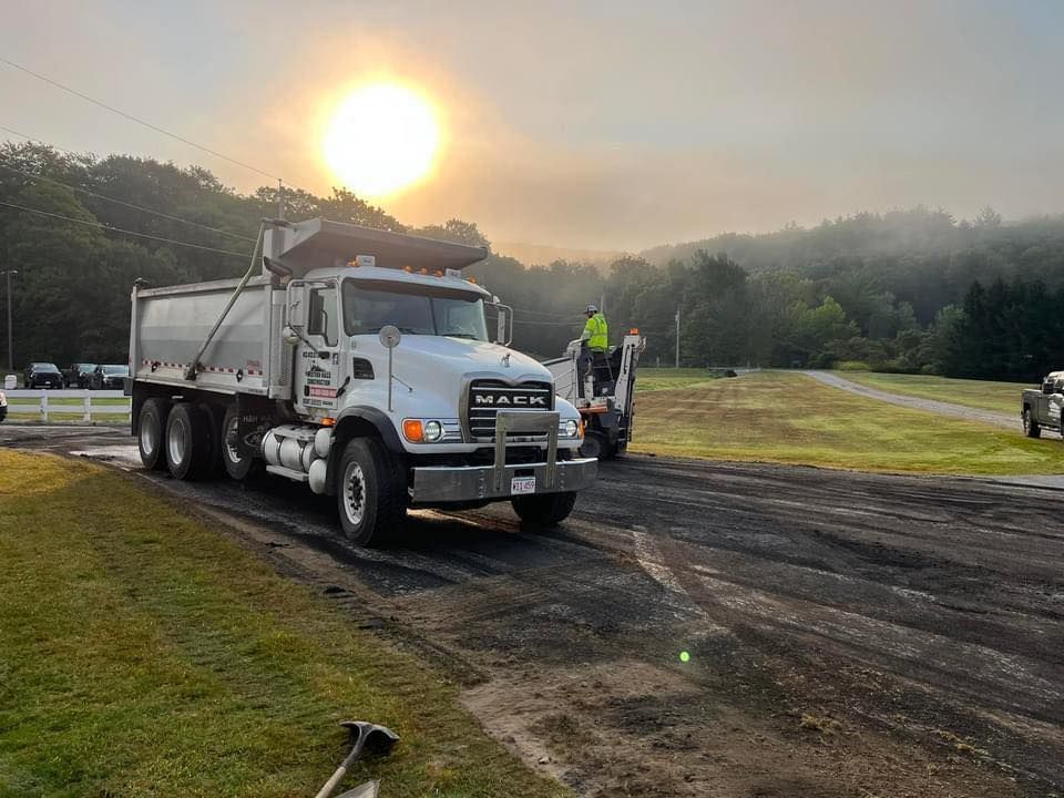 A dump truck and backhoe working on a road. Sun is rising over green hills in the background.
