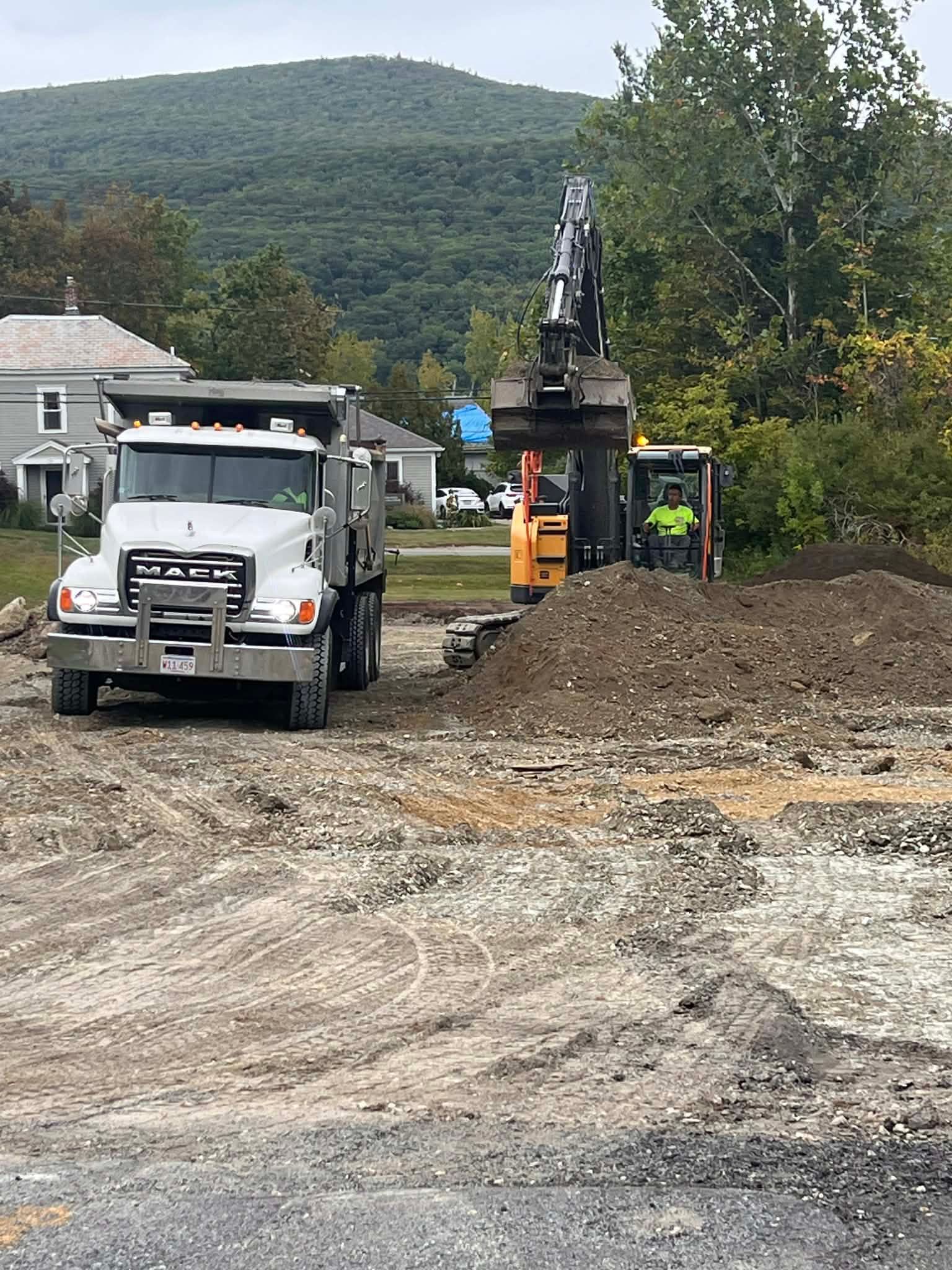 A dump truck and excavator working at a construction site. Hills and houses are in the background.