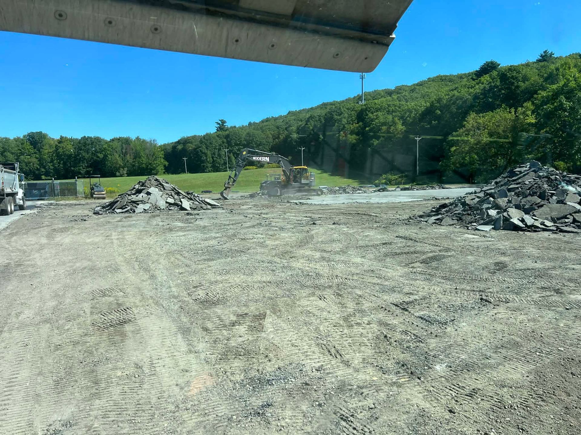 Construction site with piles of asphalt and heavy equipment; green hillside in the background.