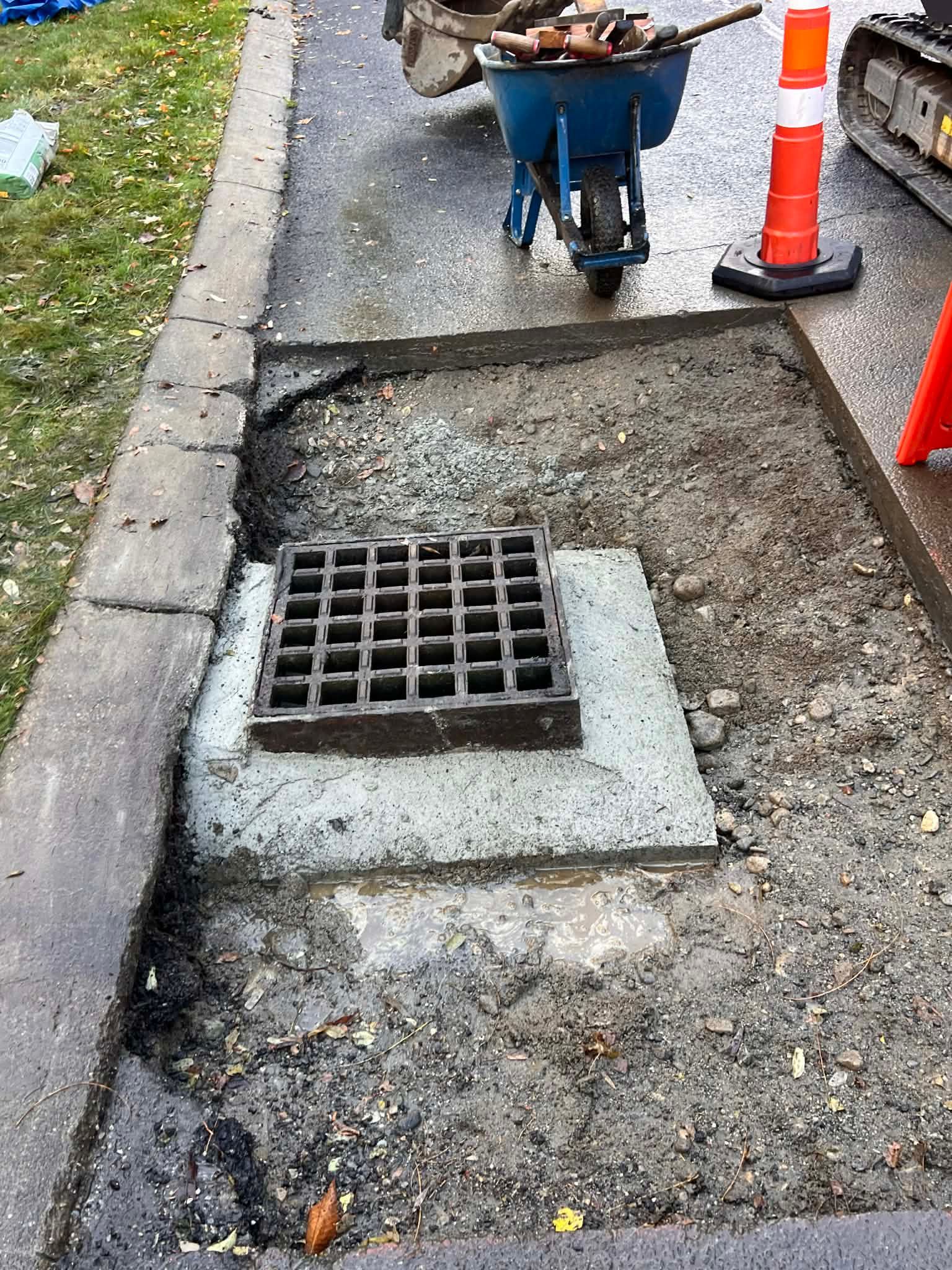 Storm drain repair in progress on a sidewalk; concrete around a grate, with a wheelbarrow and orange cones present.