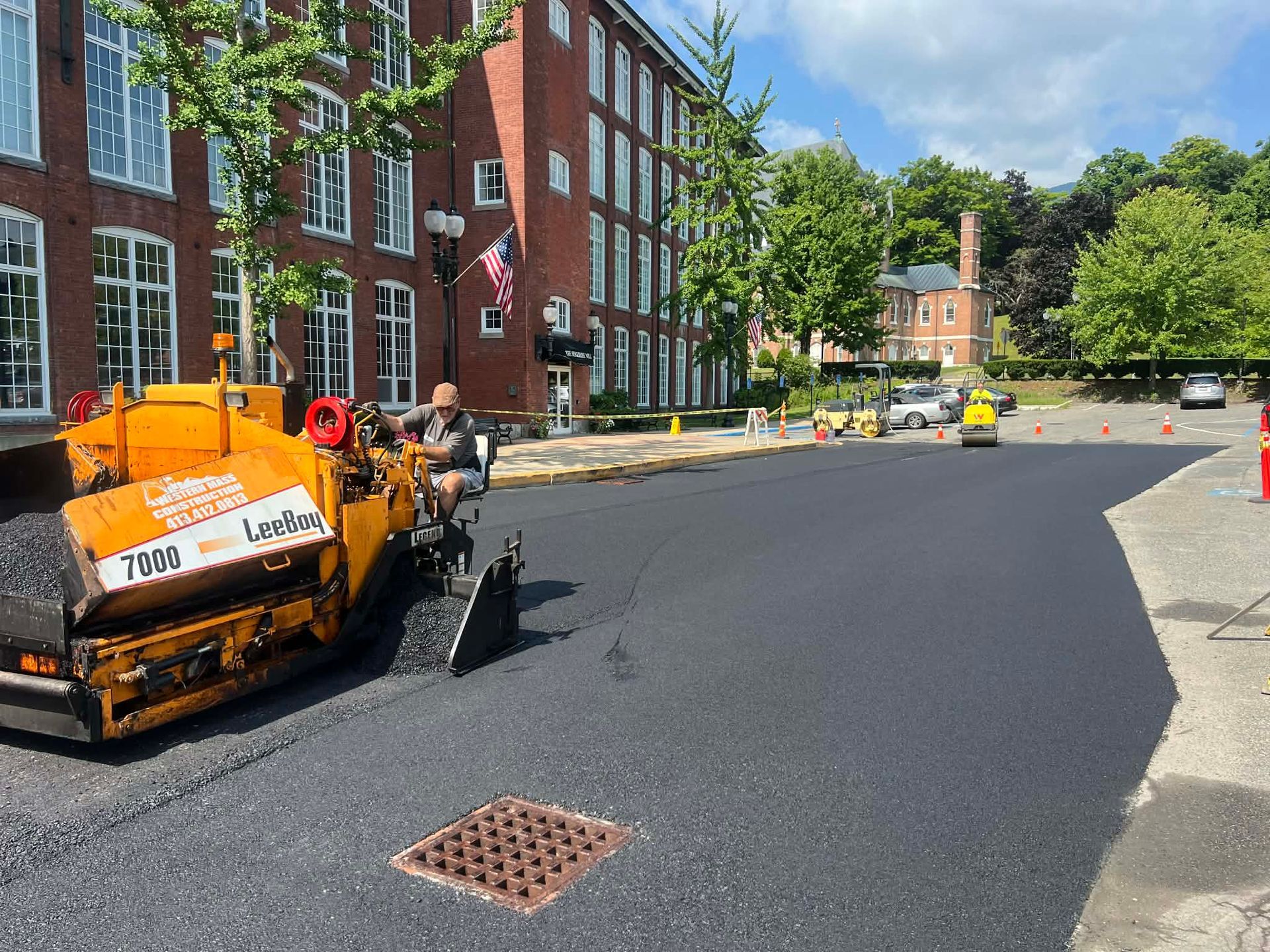 Paved black asphalt road next to a concrete sidewalk, with a red brick building and parked vehicles in the background.