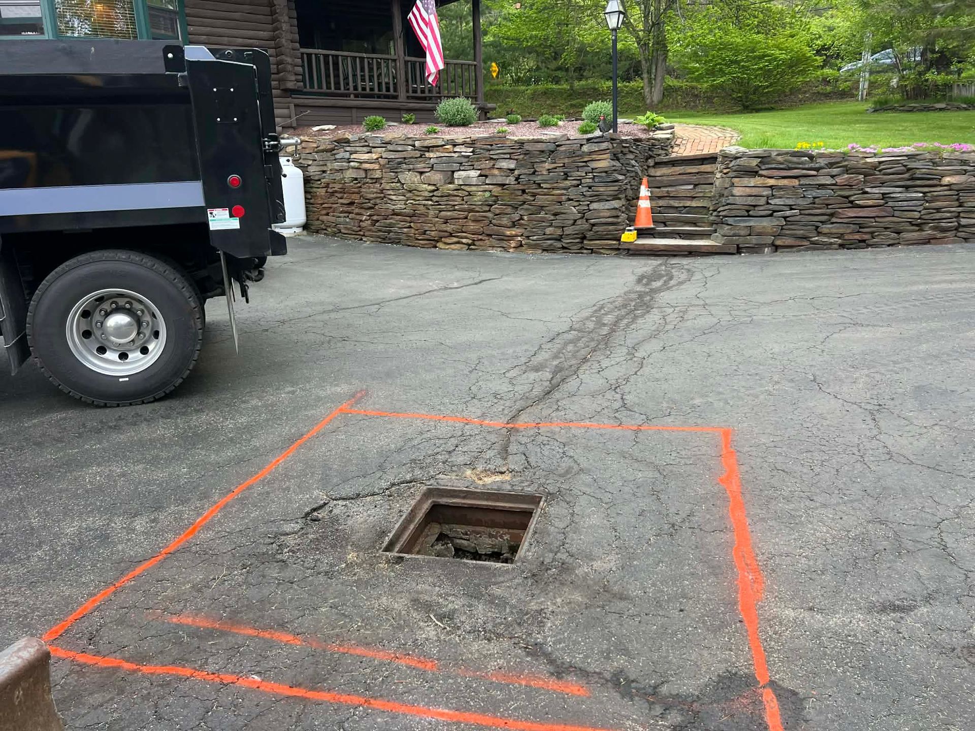 Black truck next to a rectangular opening in asphalt marked with orange paint. A stone wall and house are in the background.