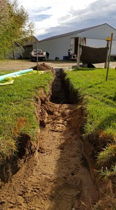 A narrow trench dug in grass leads toward a white building; a blue pipe rests nearby.