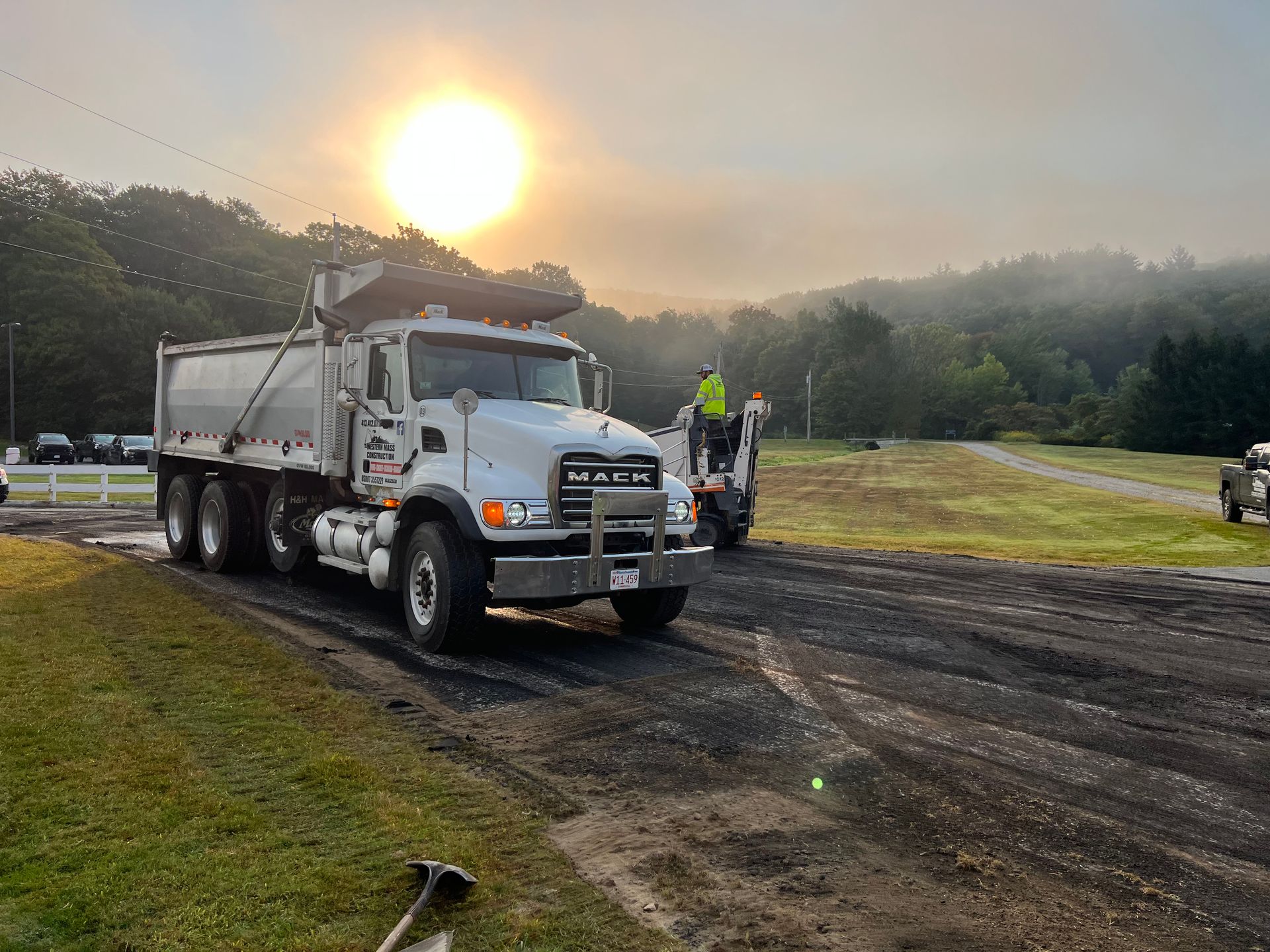 A large white dump truck parked on a dirt road. A construction worker stands near the truck, and the sun sets in the background.