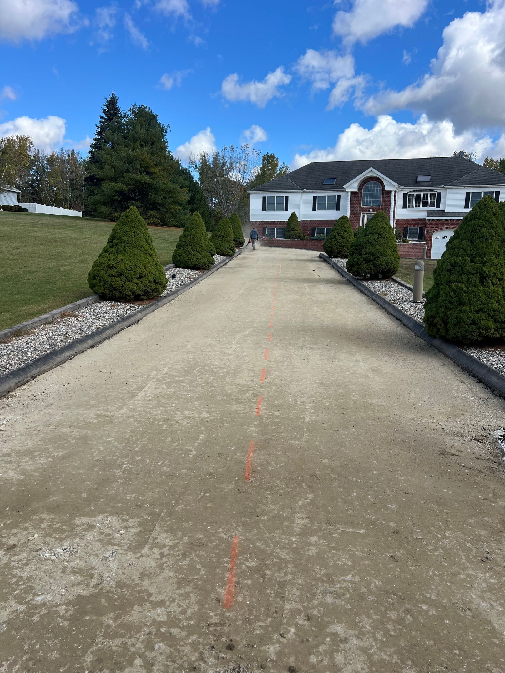 Long gravel driveway leading to a white house under a blue sky. Green shrubs line the sides.